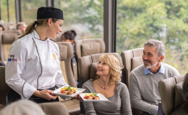 » Country Heritage Tours Happy woman and man couple in a luxury train car speaking with a dining server holding plates of food