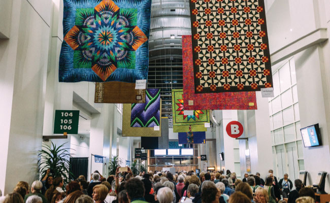 » Country Heritage Tours Conference hall with people milling about and large quilts displayed from the