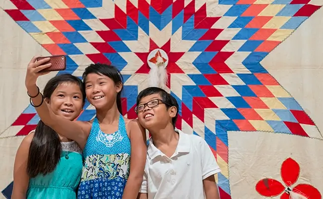 » Country Heritage Tours Three Asian children taking a selfie in front of a colorful quilt