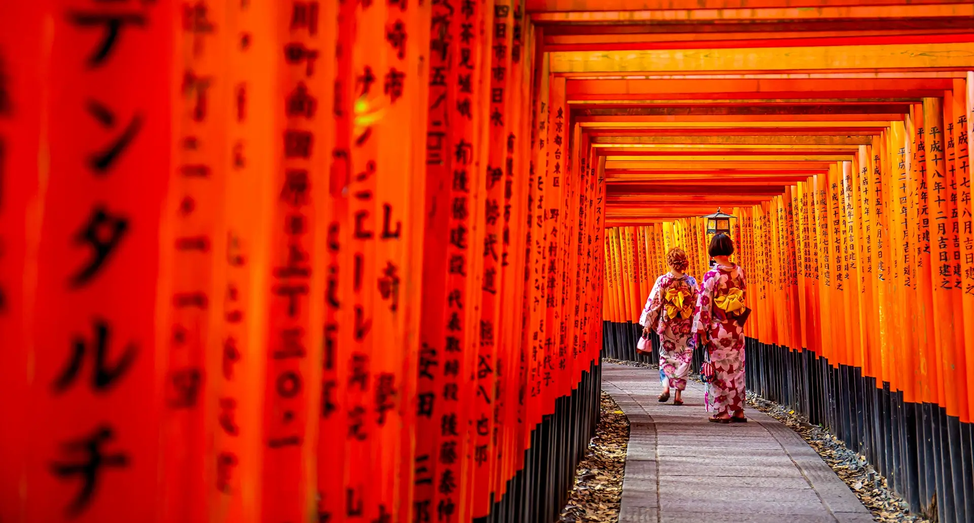 » Country Heritage Tours » Country Heritage Tours Japanese women wearing traditional clothing walking down a pathway with an archway bearing Japanese letter markings