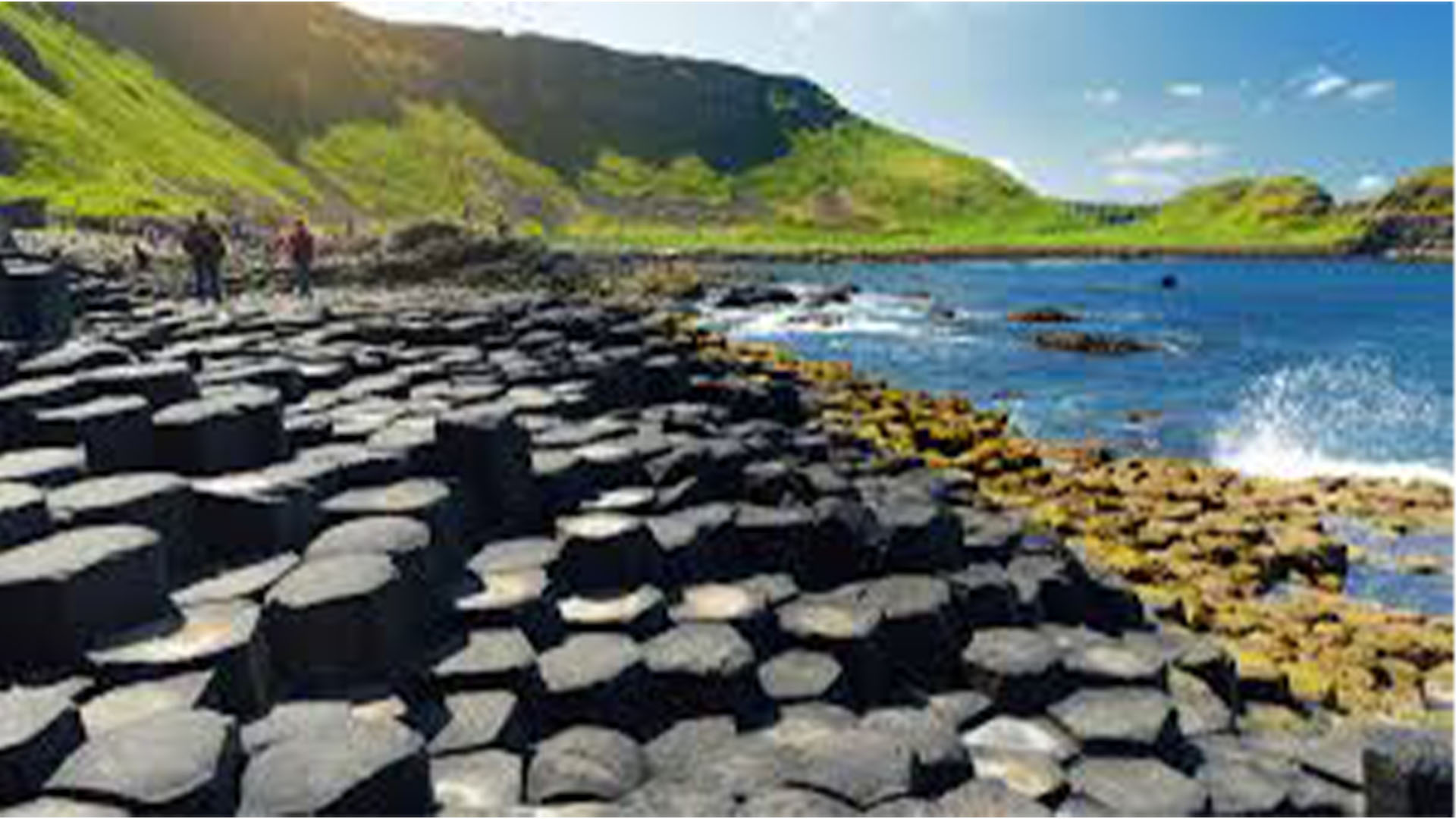» Country Heritage Tours Coastal scene with a shore of hexagonal basalt columns, blue sea, and green hills in the background; a few people walk along the rocks.