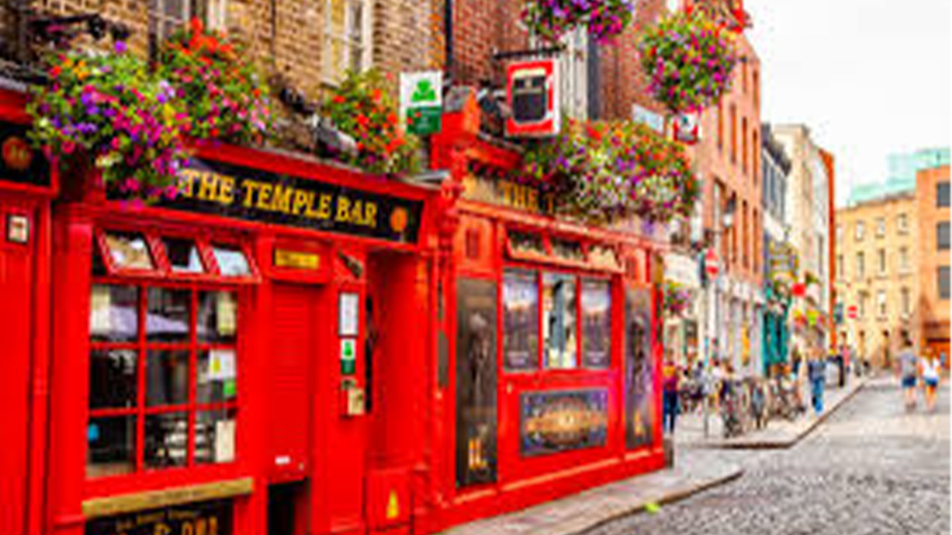 » Country Heritage Tours Bright red pub storefront with flower-filled balconies on a cobblestone street in a lively city block.