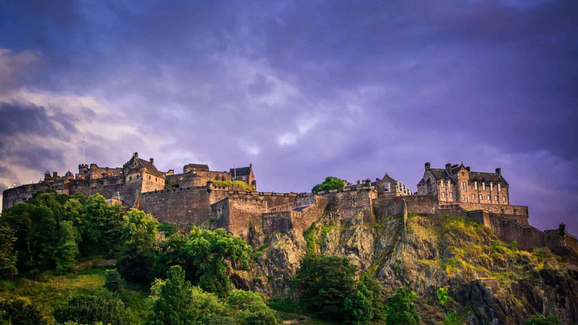 » Country Heritage Tours Castle perched on a rocky hilltop with stone walls and surrounding greenery under a purple cloudy sky.